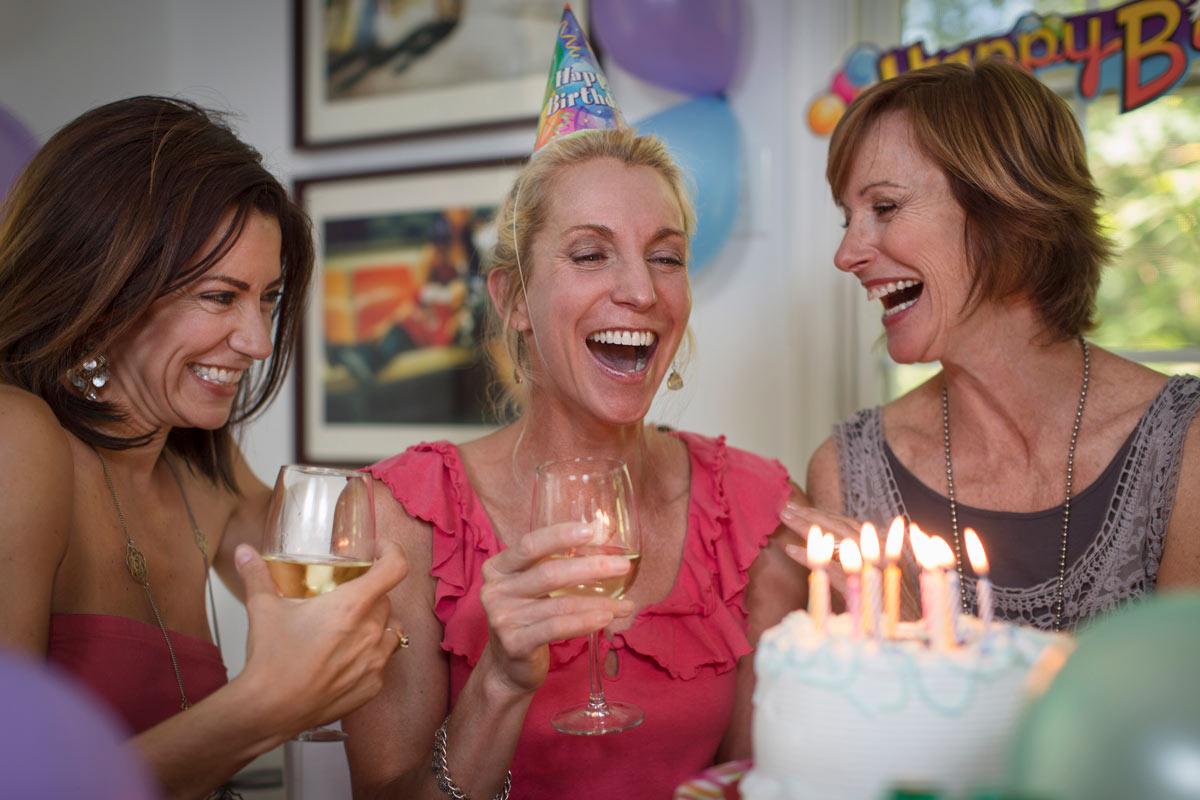 Three white 55-year-old women with wine glasses laughing at a birthday party in front of a cake with lit candles.