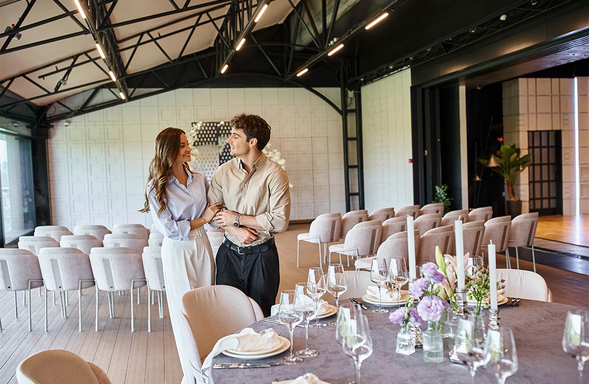 white man and woman in their late 20s lock arms and walk and talk through a wedding venue set up with chairs and dinner tables indoors 