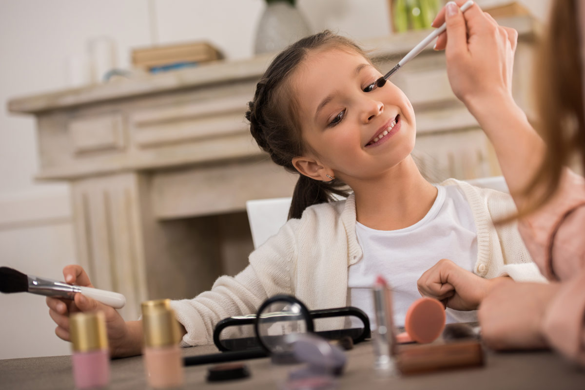 A person applying makeup to a smiling white girl as makeup sits scattered on the table in front of them.