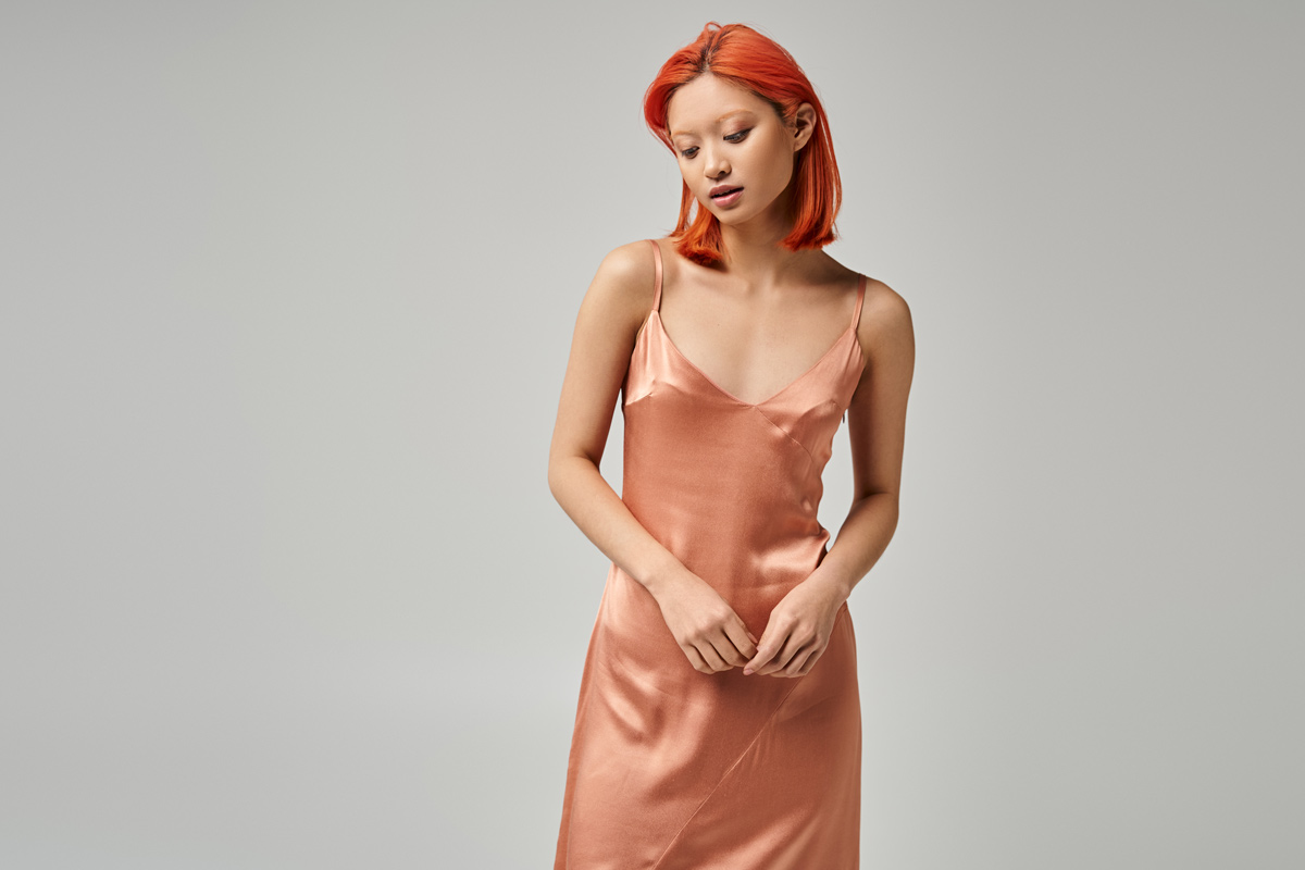 Asian 25-year-old woman with orange hair looking down while wearing a pink satin dress.