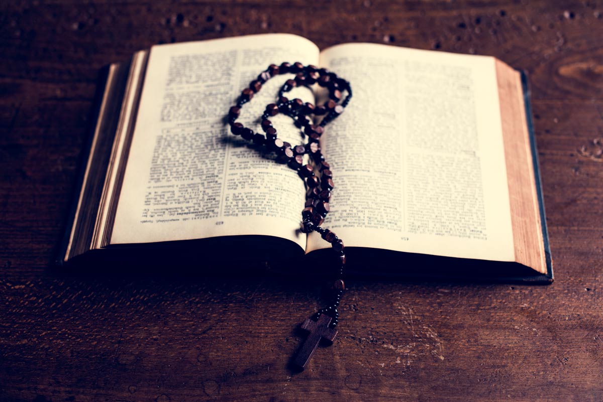 Bible laid open on a dark wooden desk with rosary bead necklace placed in between pages