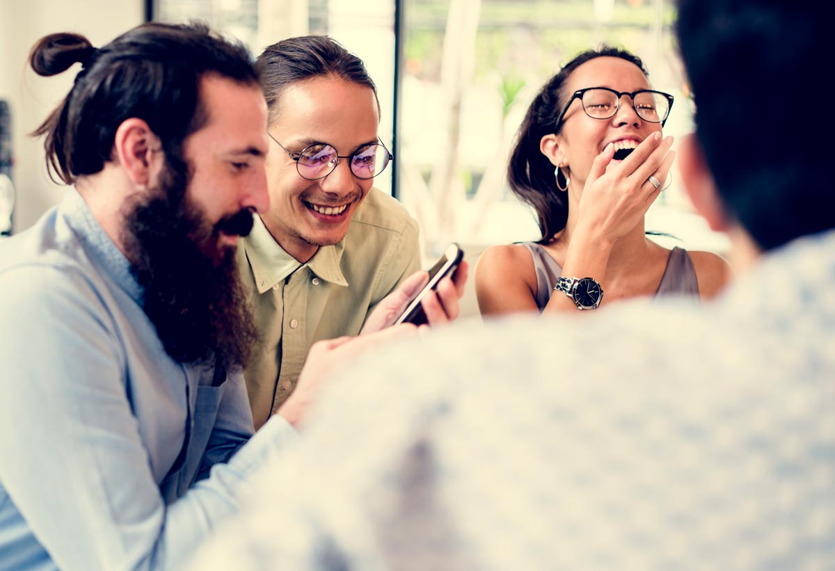 A group of 4 white middle-aged friends sit together, laughing while watching something on their phones. 