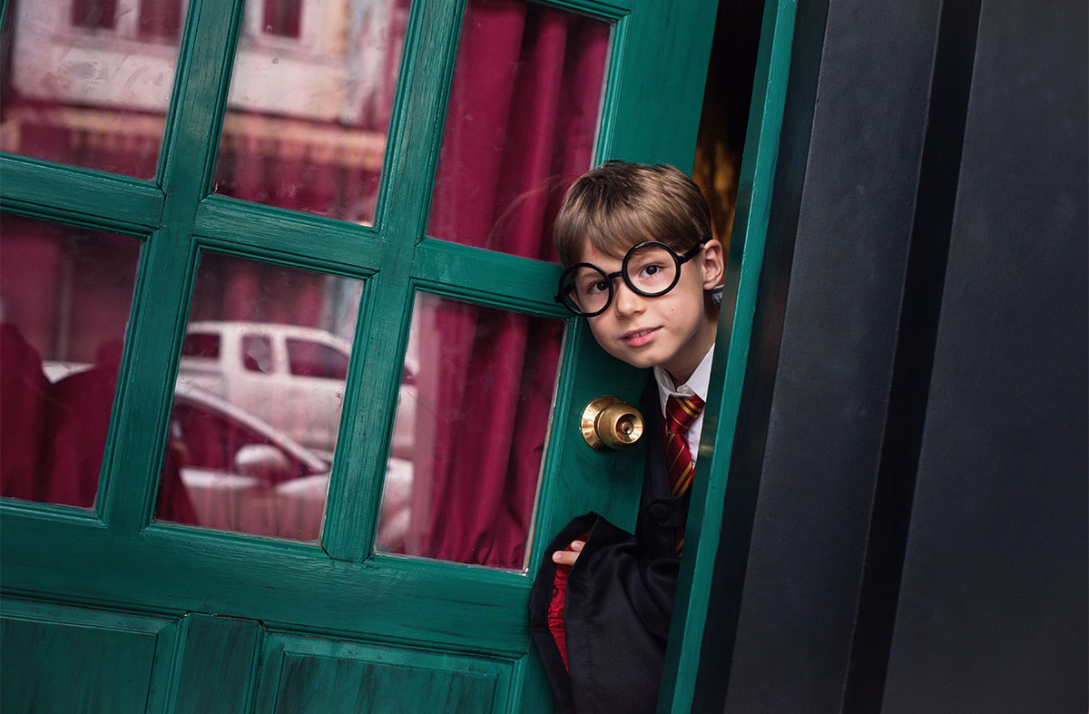 White little boy, wearing glasses and a wizard costume, poking his head through a green door. 