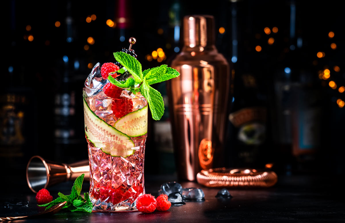 Raspberries, mint, and cucumbers in a cocktail glass, with red liquid in them, on a black table with a background of black with other cocktail supplies. 