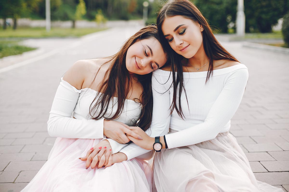 Two white brunette sisters in their early 20s wearing matching white long sleeves hold hands and lean heads together while outside on a path