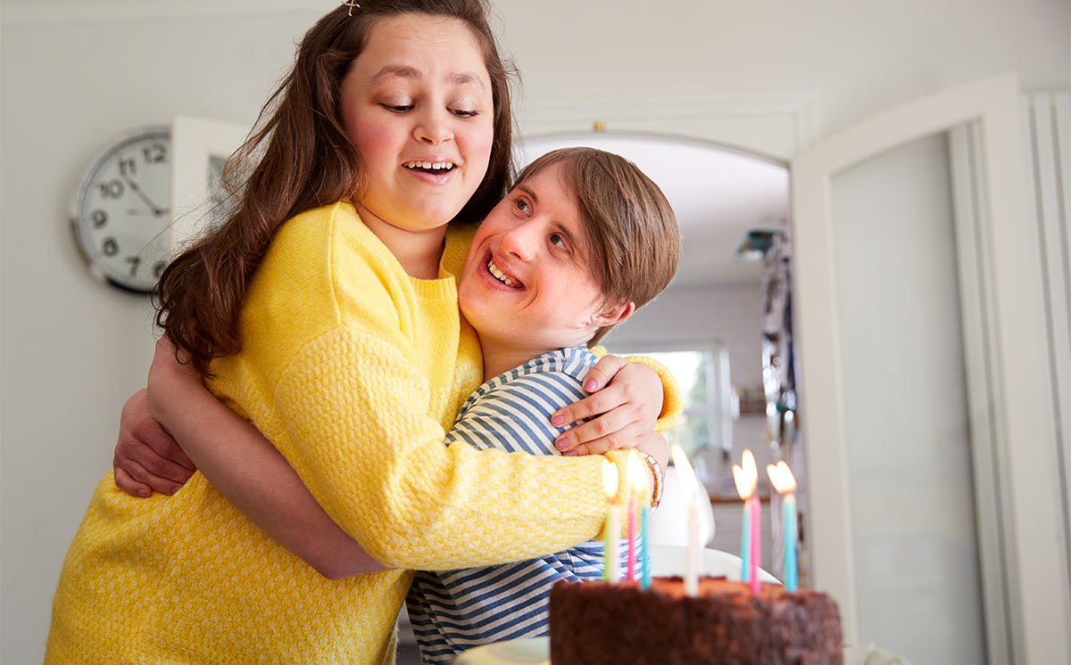 Two white teens hug indoors over birthday cake with lit candles