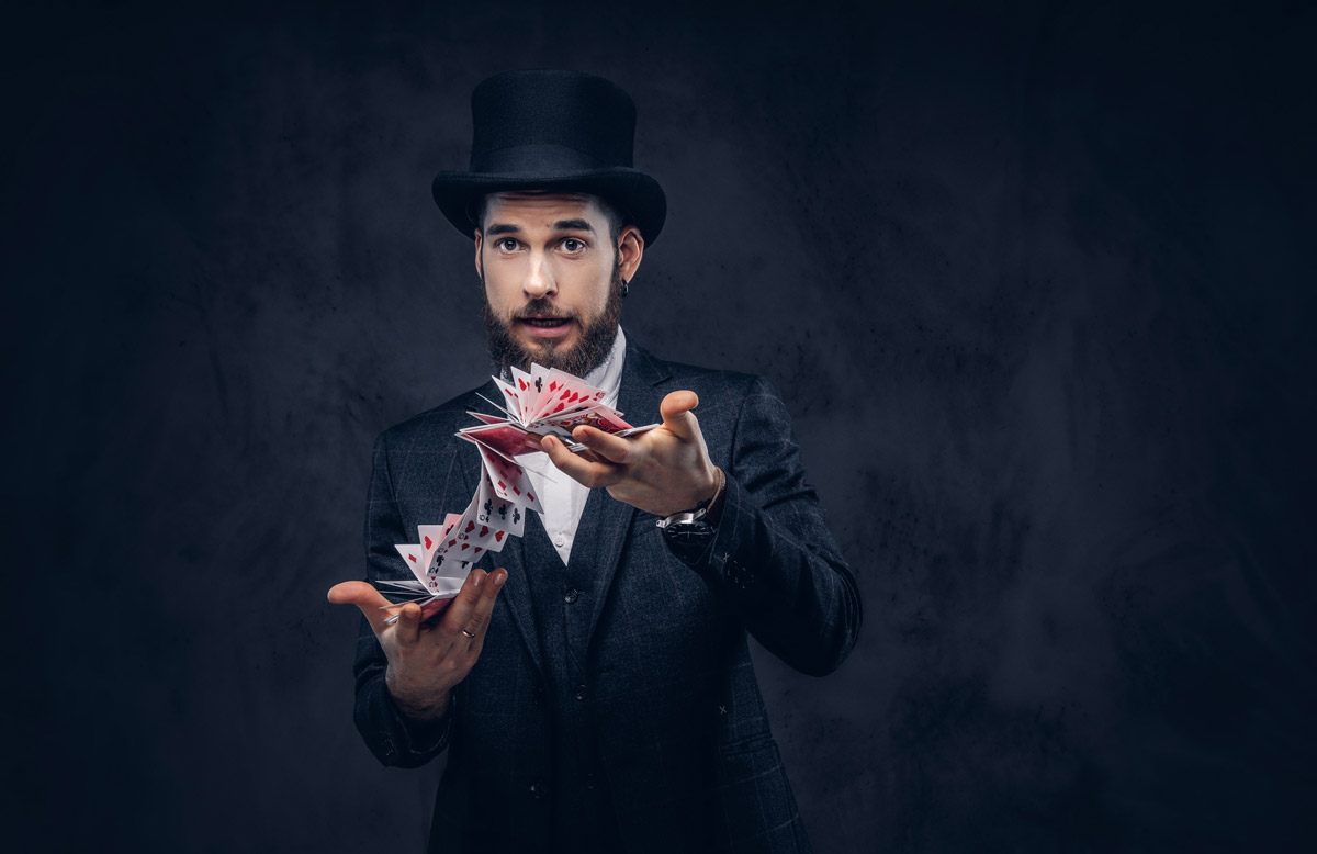 A white man with a black beard wearing a black top hat and a suit flourishes a deck of cards.