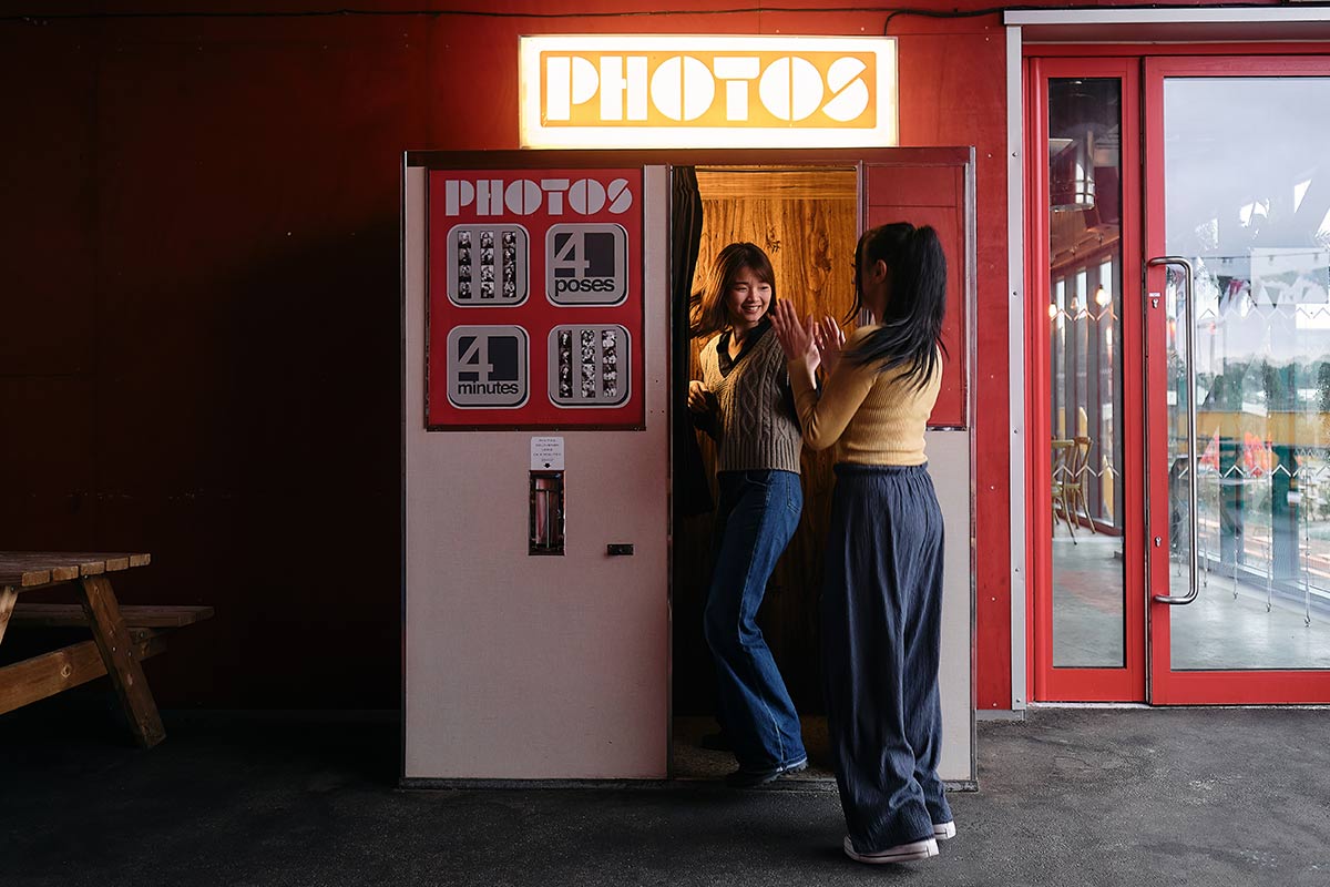 Two 24-year-old asian women smiling enter a vintage photo booth in a mall.