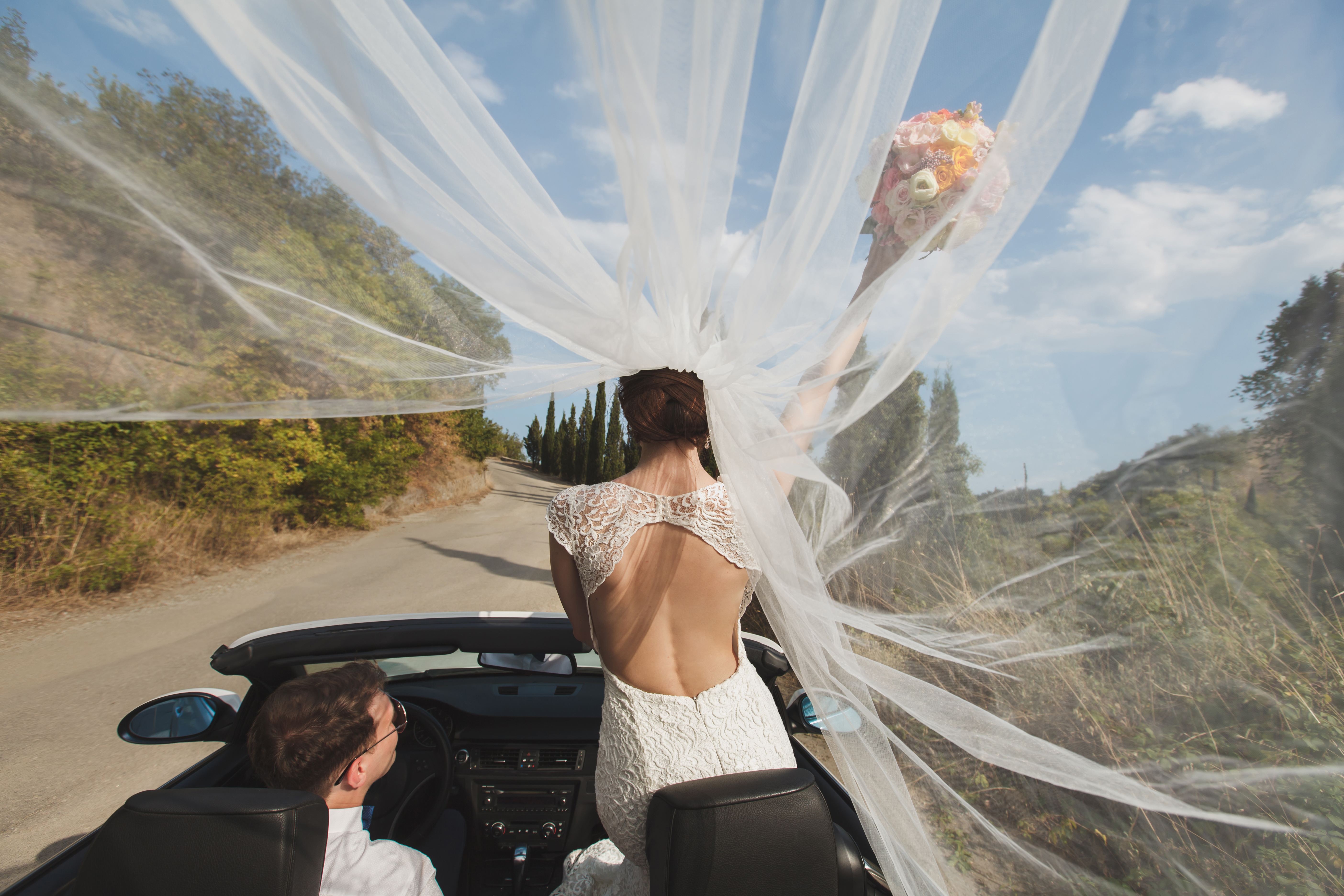 White couple in their twenties driving wedding convertable getaway car while the women stands in the passanger seat holding her bouquet of flowers above her head and her veil blowing behind her in the wind