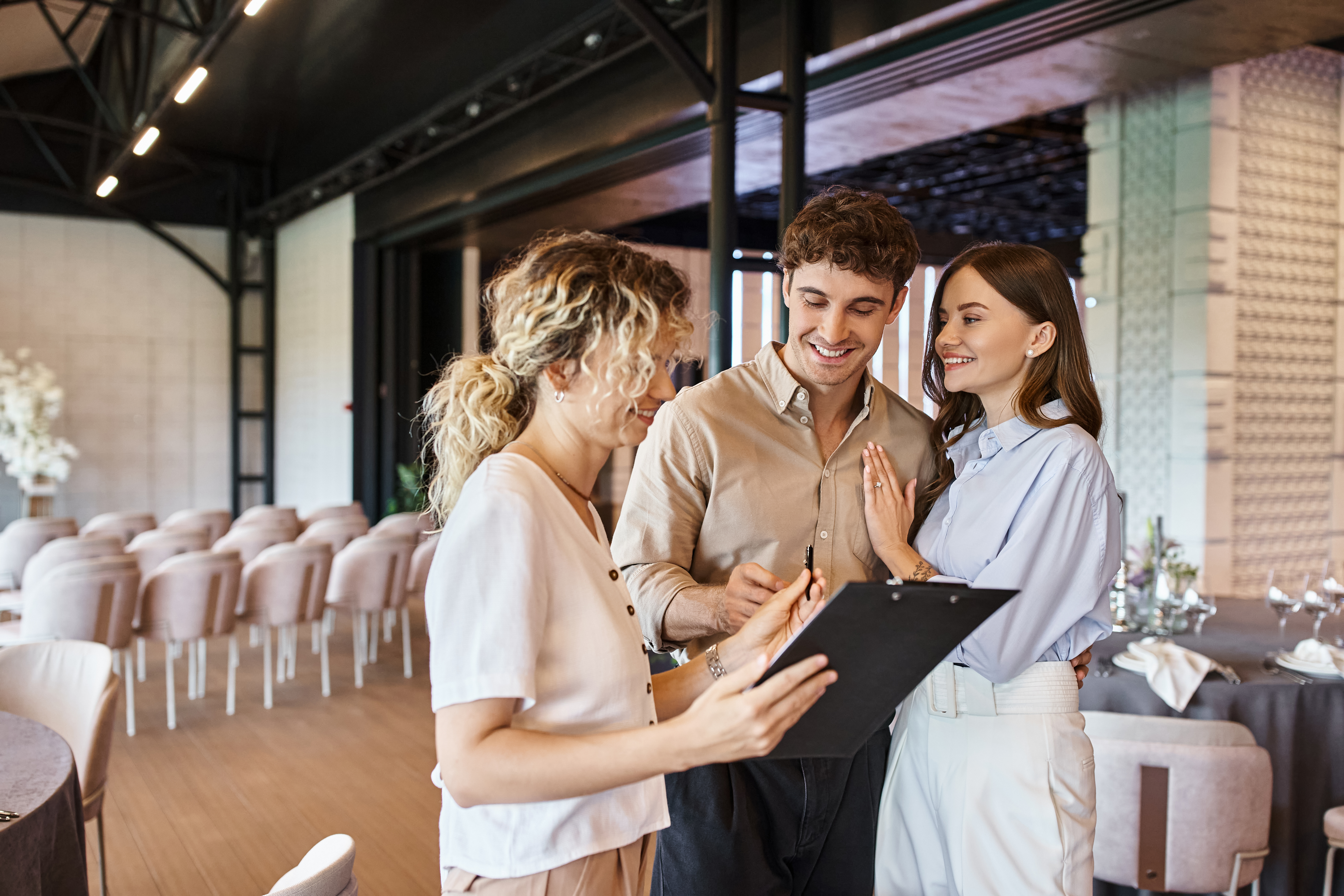  White 20 something man and woman smile and speak with 30 something white woman holding clipboard in an indoor wedding reception venue setting 