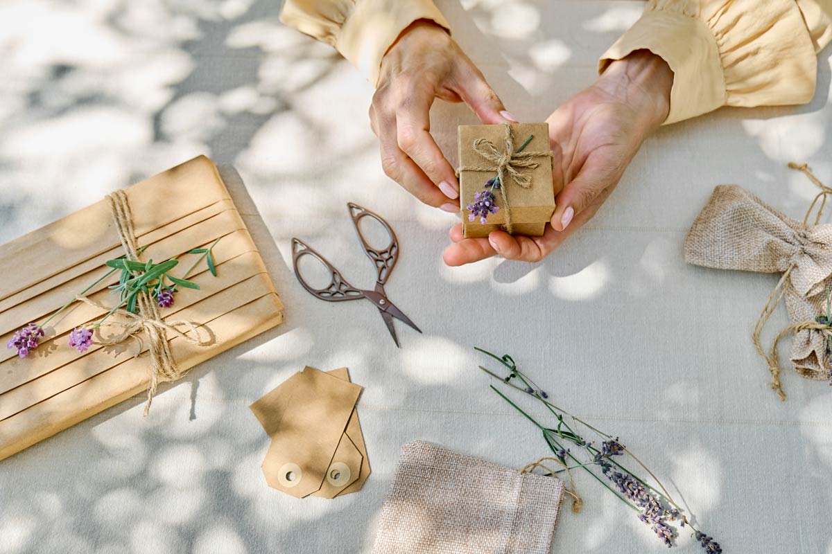 Woman in yellow long sleeve blouse wrapping a handmade gift with craft paper and a twine on a white table with silver small scissors, lavender, and brown paper gift tags