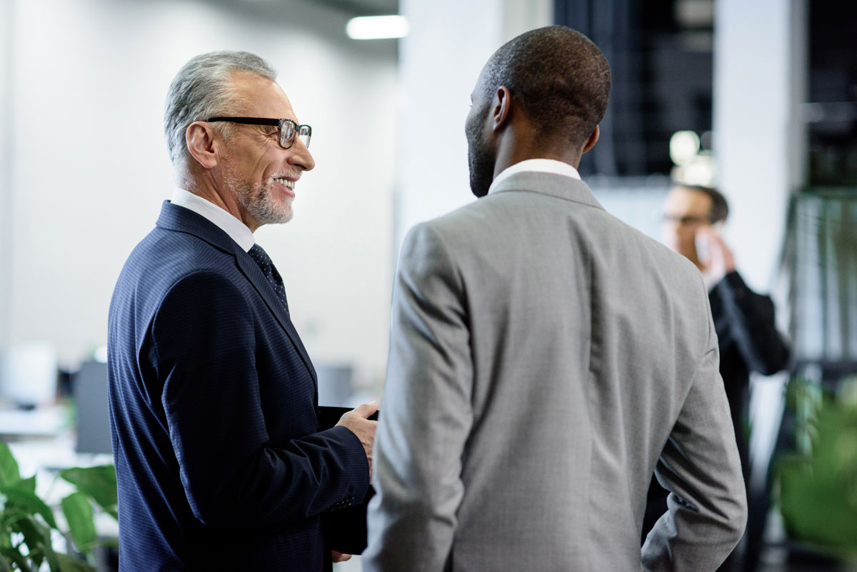 One older white man, and an older black man stand in suits conversing in a business setting.