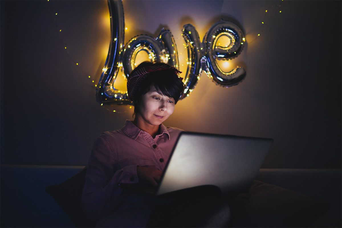 30 something women stares at computer in the dark with light up love sign on the wall behind her