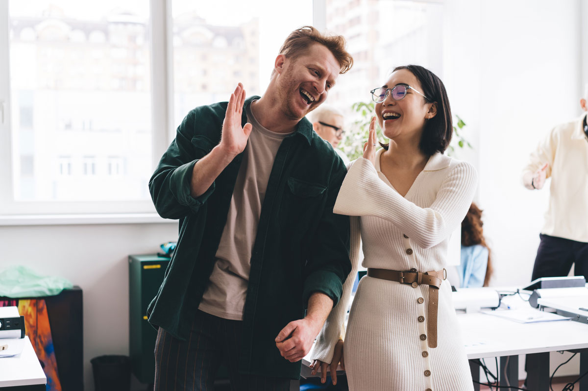 A happy white man and woman in their 30s in a bright office setting share a celebratory high-five.