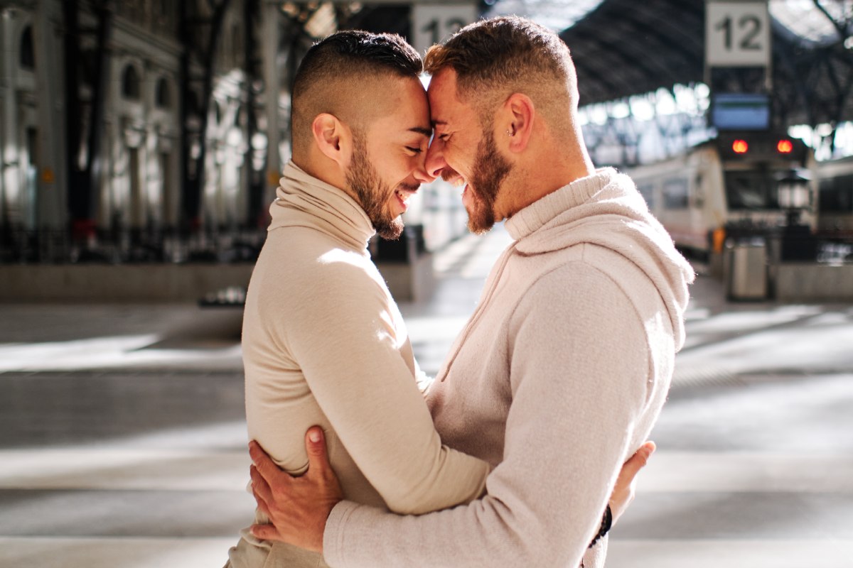 30-something white gay couple in neutral colors embracing each other, smiling near a subway outside