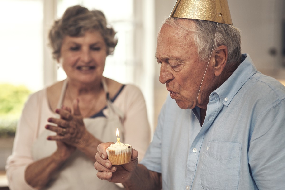 white elderly man blowing out a candle on a cupcake with a golden birthday hat as an elderly woman is clapping for him in the background smiling with an apron on