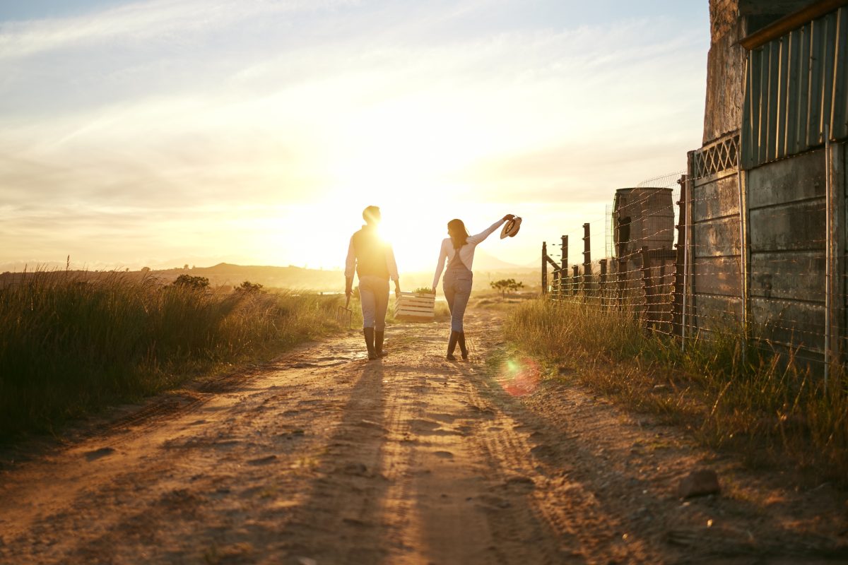 couple walking towards the sunset on a farm holding a basket together with a hat hanging on the woman's arm raised