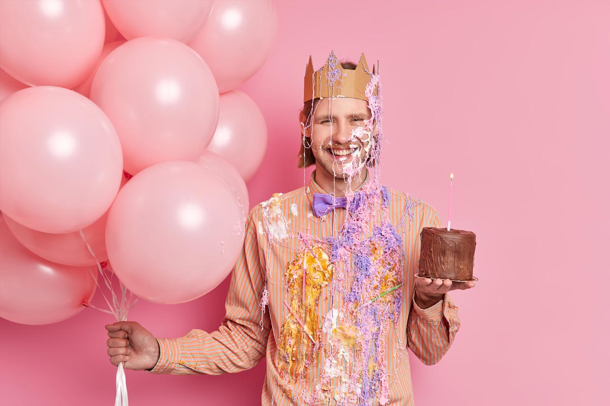 A happy white 20 something man in a crown stands against a pink background, holding a small chocolate cake in one hand and a bunch of pink balloons in the other, his shirt and face smeared with cake and frosting.