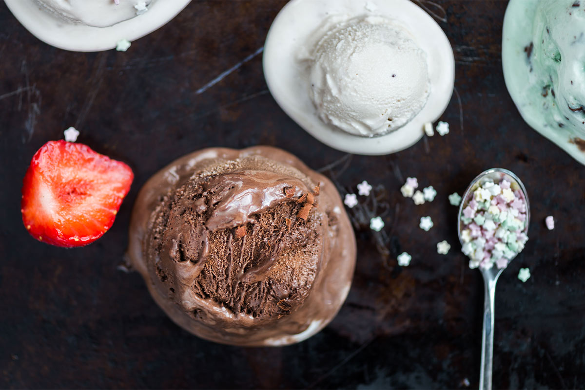 scoops of chocolate, vanilla, and mint chocolate chip ice cream sit on a black surface next to a strawberry and a serving spoon with small star sprinkles on it