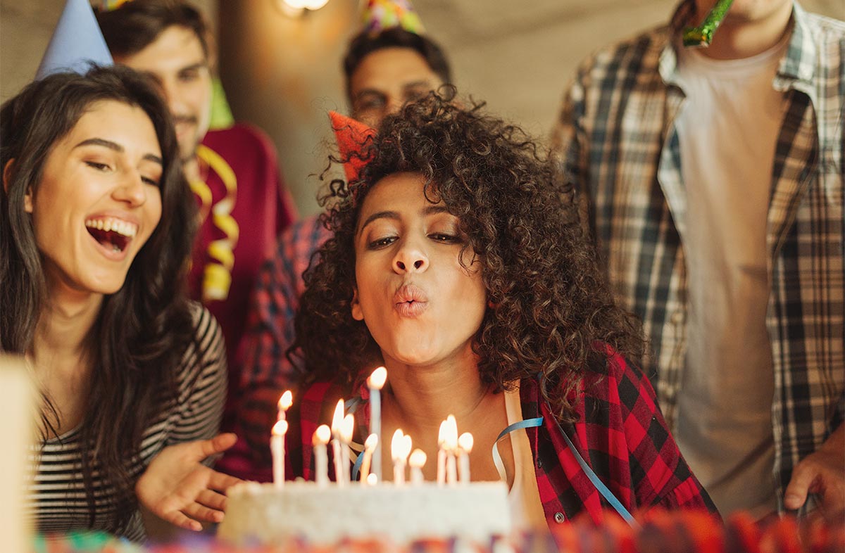 30-something black woman blows out birthday candles while adult friends cheer in party hats around her