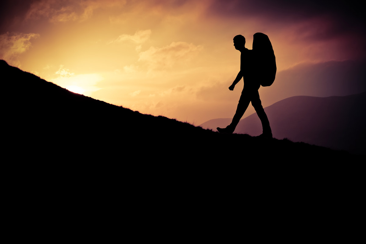 A silhouette of a man with a large backpack hiking up a hill in front of a sunrise.