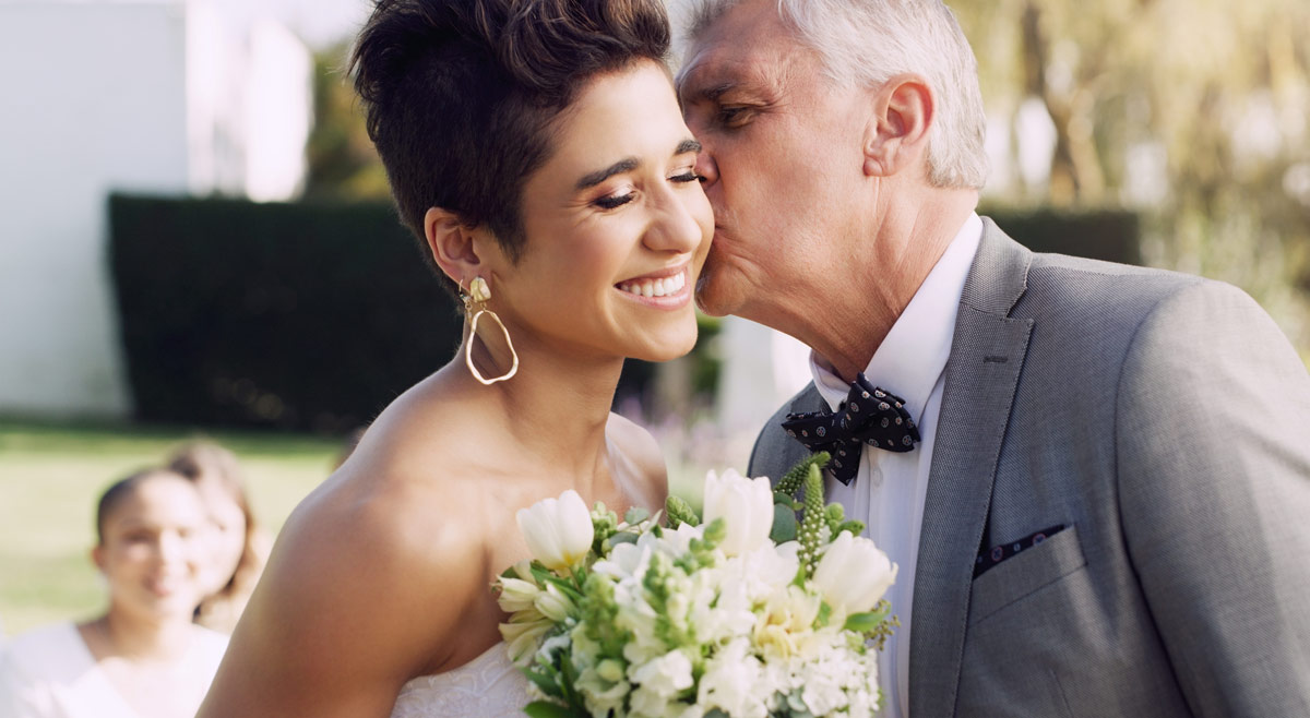 bride with short hair smiling and holding bouquet as her well-dressed older father kisses her cheek