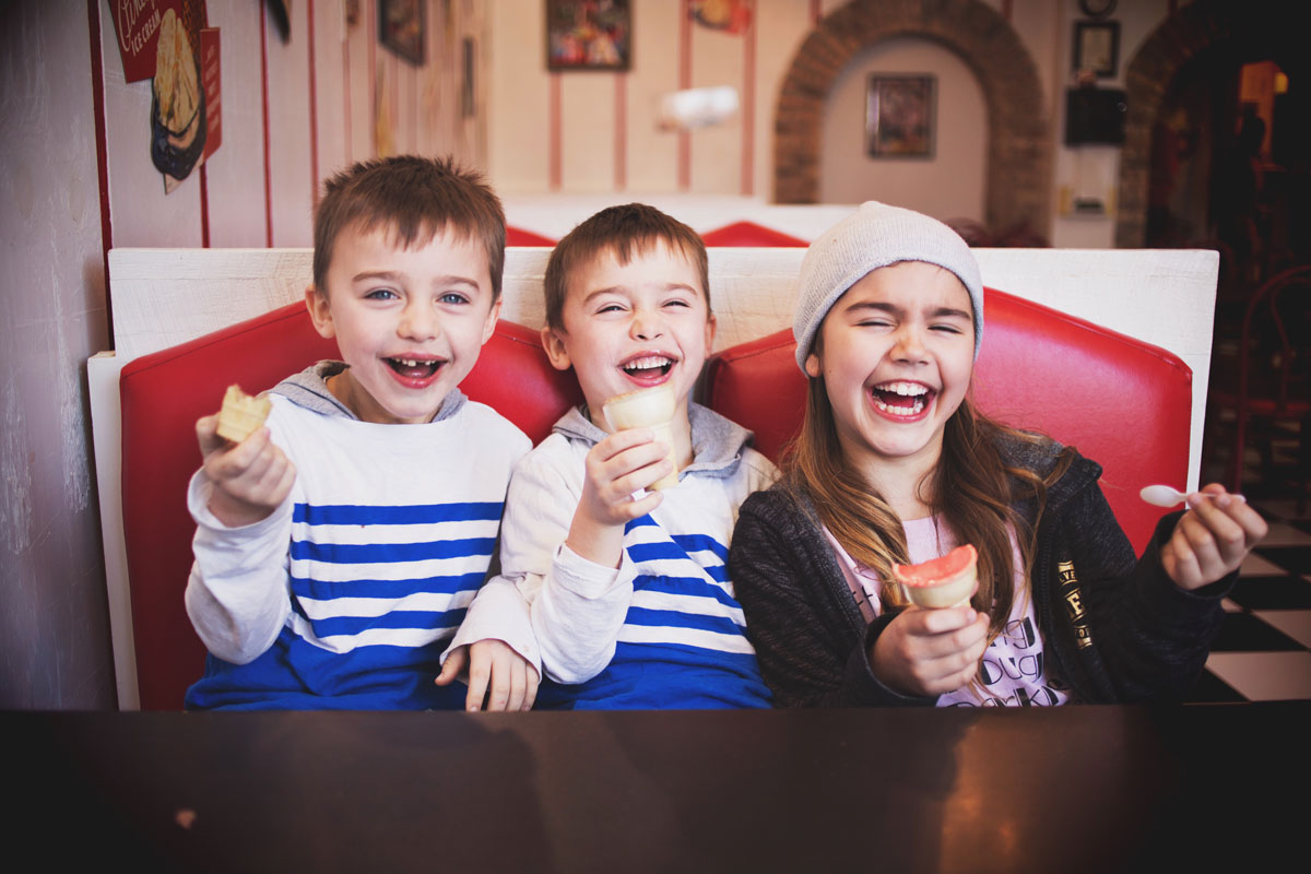 Three young white kids age 7-9 enjoying ice cream and laughing in a booth at indoor ice cream parlor