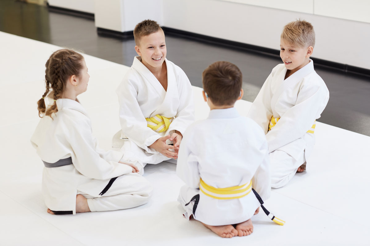 Four white kids age 10-12 in karate attire and yellow and gray belts sit on knees and talk on a karate floor mat inside