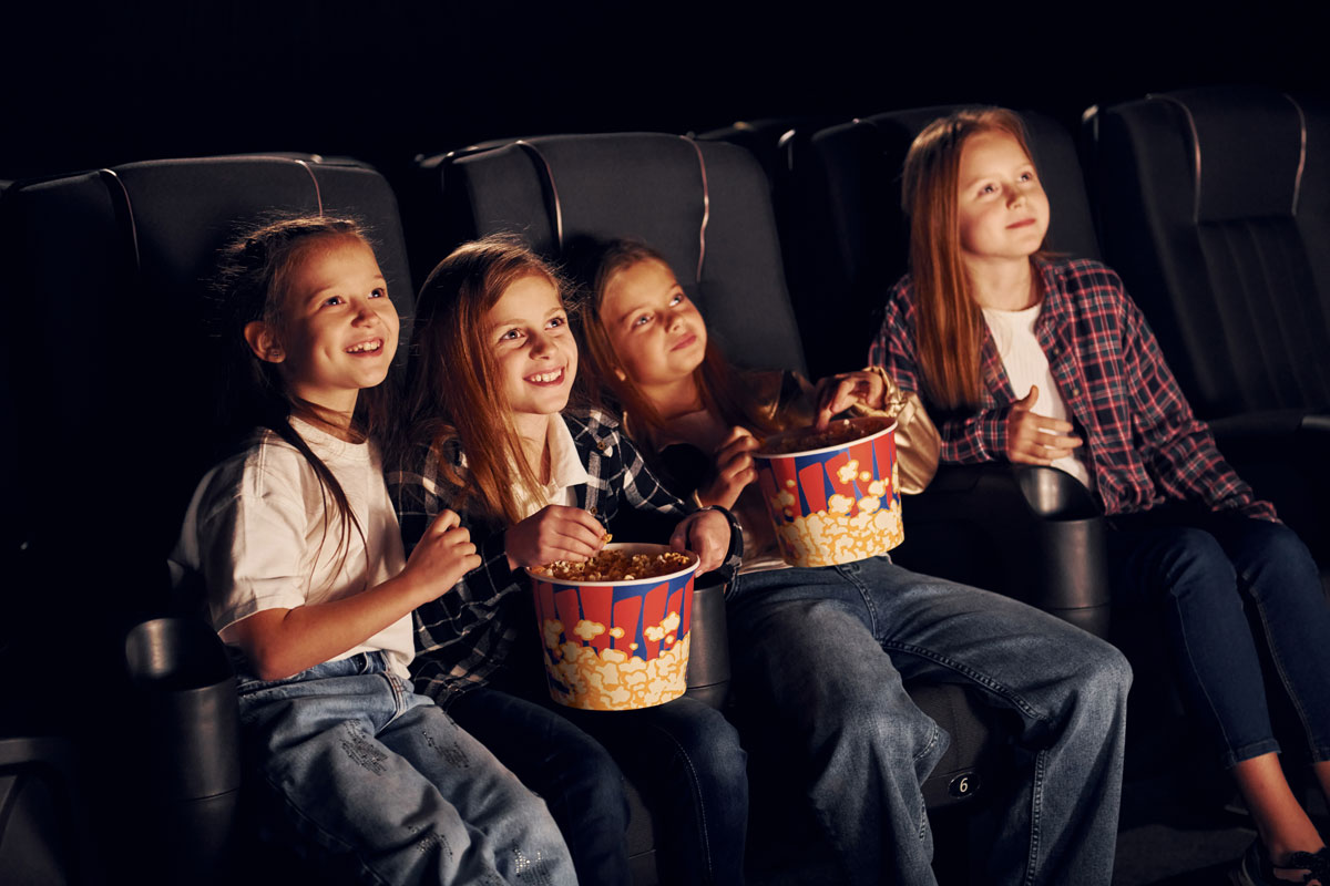 Four young white girls ages 8 to 10 smile while watching a movie in theater with popcorn buckets