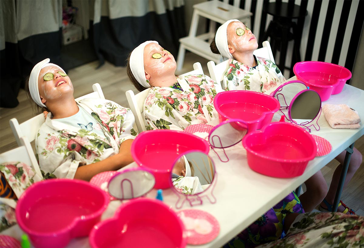 Young white girls enjoying face masks and cucumbers on their eyes while sitting back in floral robes and white headbands at a diy pink and white spa table