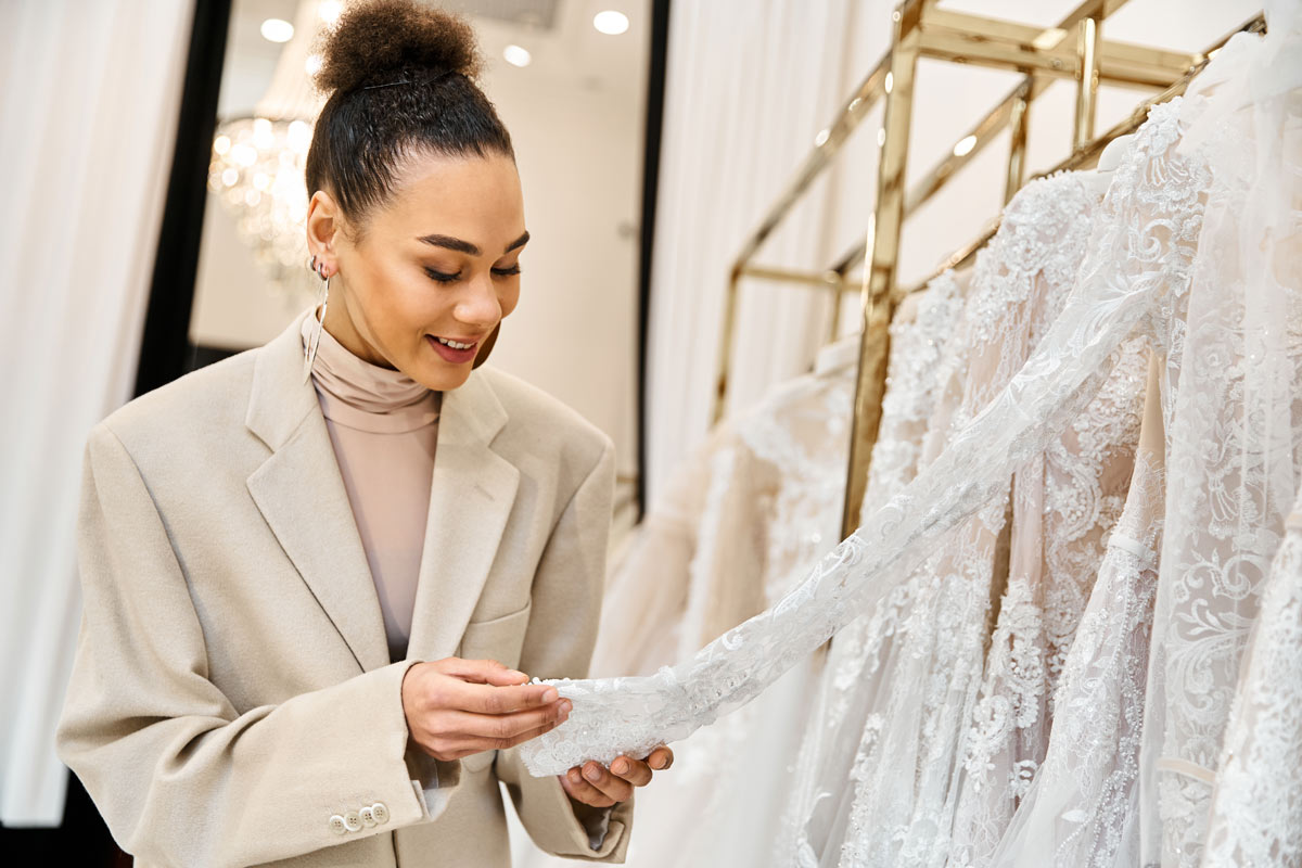 Mixed-race 30-year-old woman wearing a beige blazer looking at lace wedding dresses in a bridal shop. 