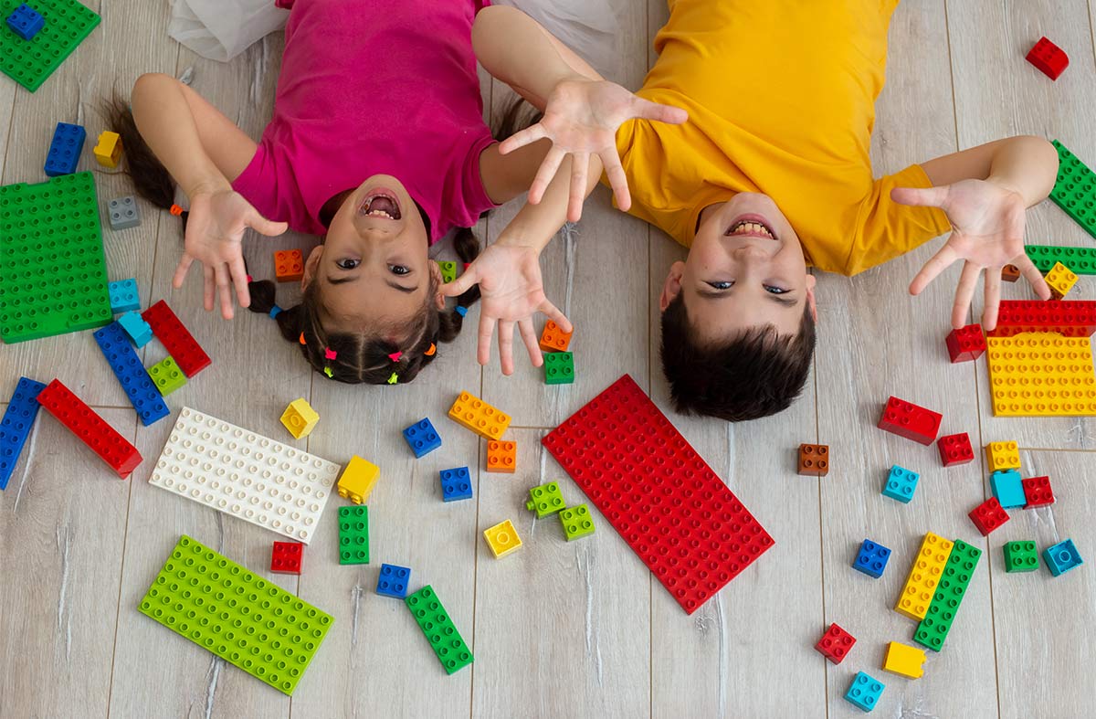 Little boy and girl laying down on white wooden floor, with legos around them. With little kids hands in the air