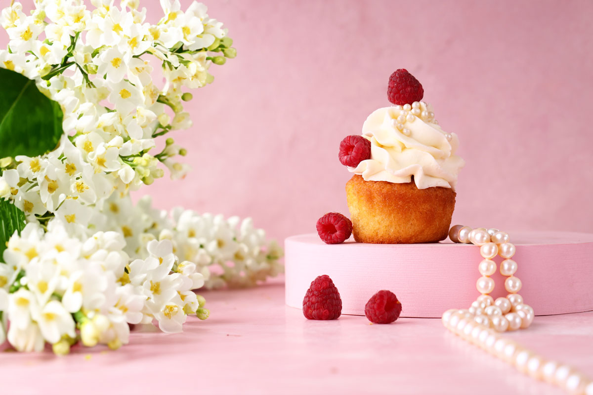 Vanilla cupcake with raspberries sits by pearls and white flowers on a pink backdrop