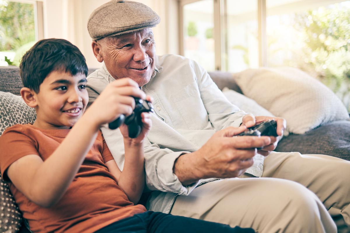 Indian grandpa and 8-year-old grandson holding video game controllers playing video games on the couch smiling 