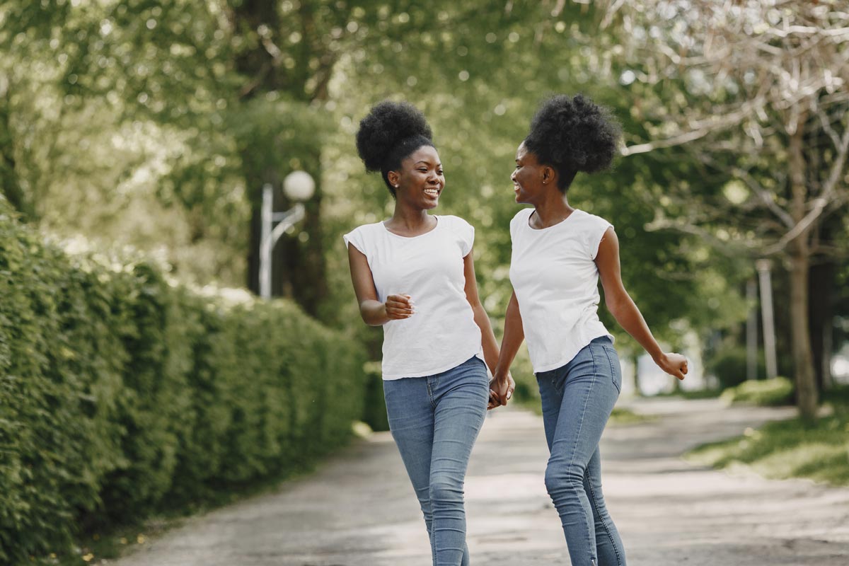 Two black sisters in their late teens wearing identical bun, white shirt, and jeans, hold hands and talk as they walk down a sidewalk path by bushes and trees 