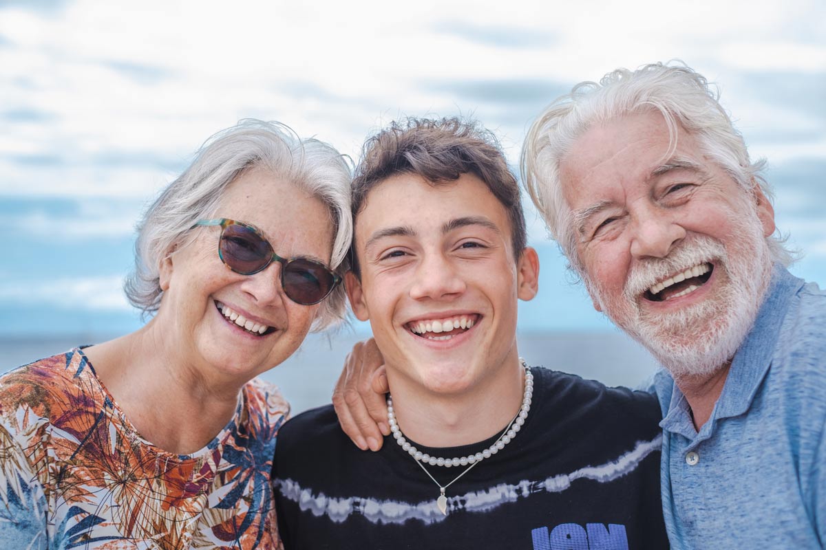 white grandparents smiling with their teen grandson outside with sunny blue skies side by side