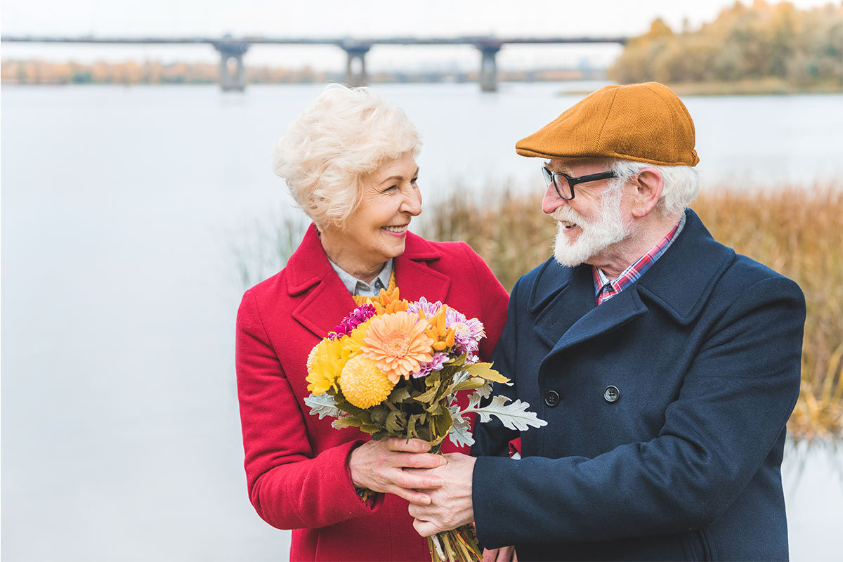 Elderly white man hands flowers to elderly white wife at a lake