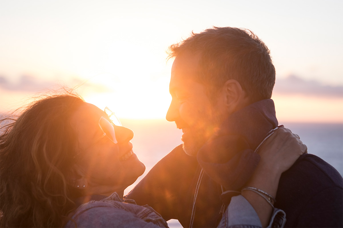 White young adult couple smiling and embracing as the sun shines brightly behind them