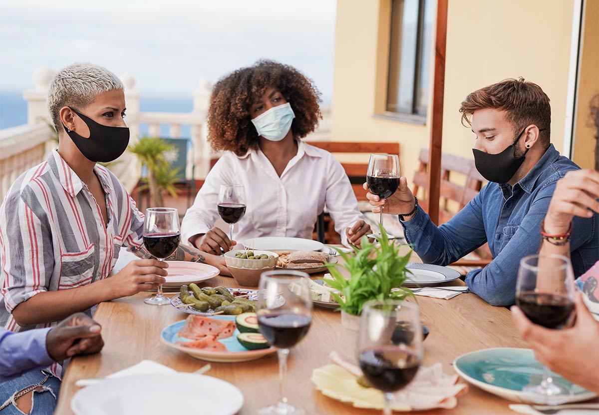 several people of multiple races wearing protective face masks sit at an outdoor dinner table that has glasses of wine and miscellaneous foods on plates