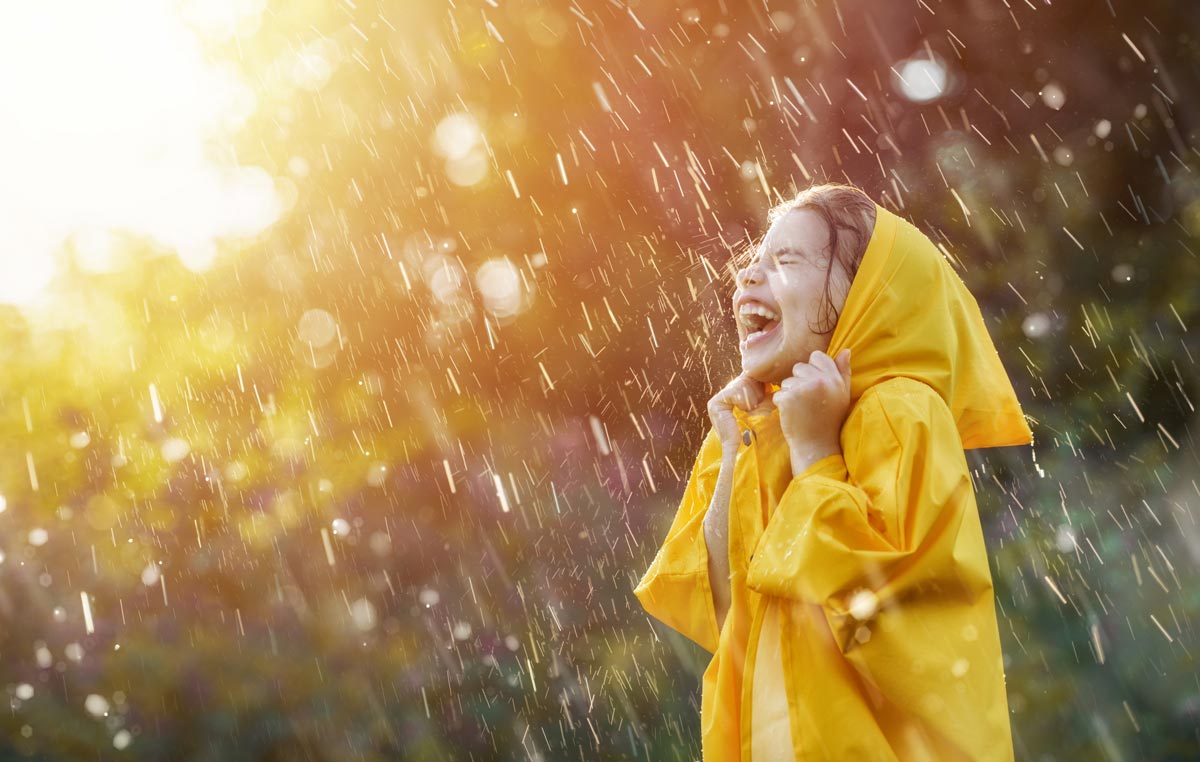 A little white girl wearing a yellow raincoat laughing while playing in a sunny rain storm