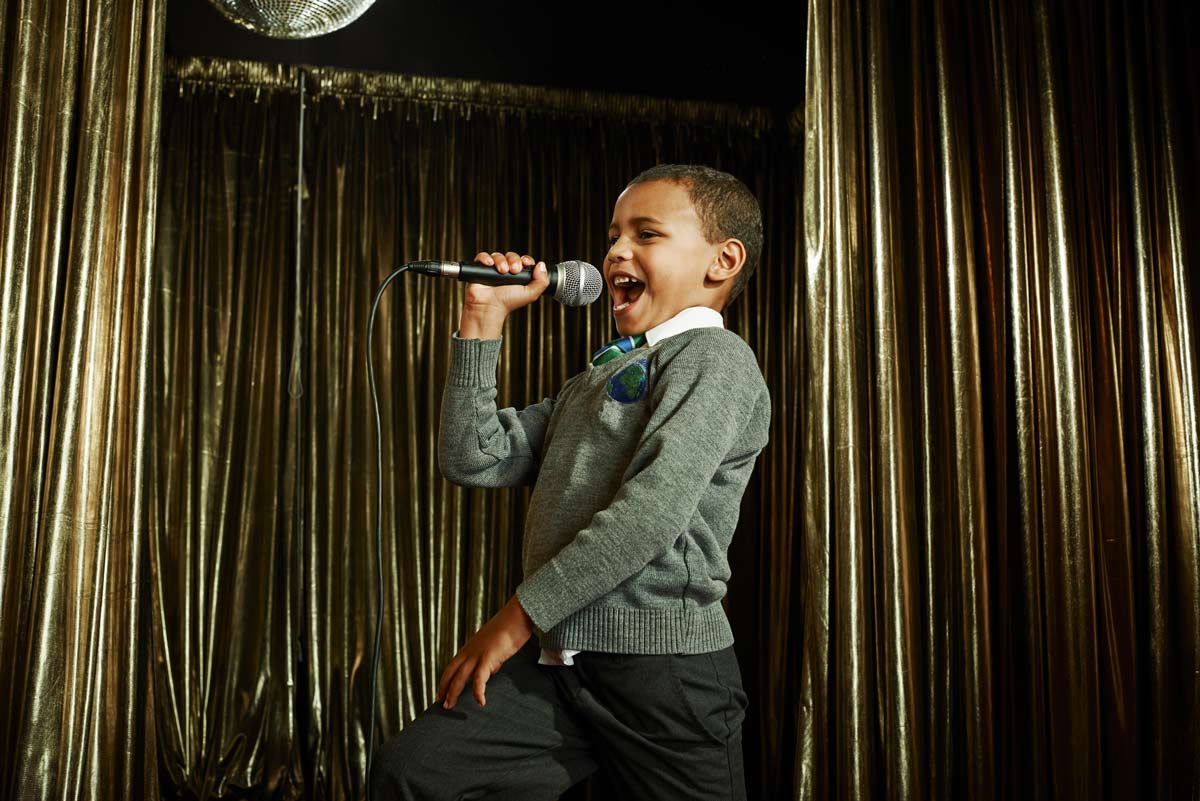 5-year-old mixed boy singing in a microphone with golden metallic drapes and a disco ball above him dressed in a sweater and a collard shirt with a tie