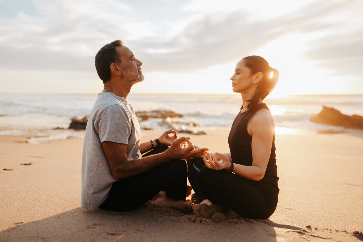 50 something white couple meditate on the beach with their eyes closed while the waves wash in and the sun shines behind them.