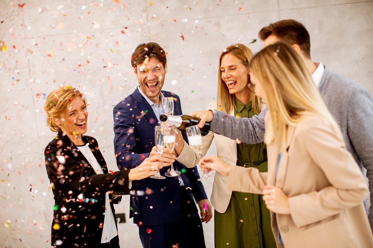 Group of 5 white middle-aged coworkers, wearing blazers, sharing champagne and smiling. 