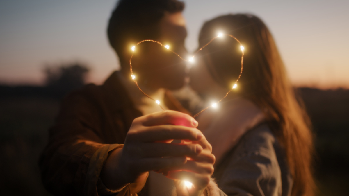 a 20 something white couple kiss and hold up a light up heart outdoors while the sun sets behind them.