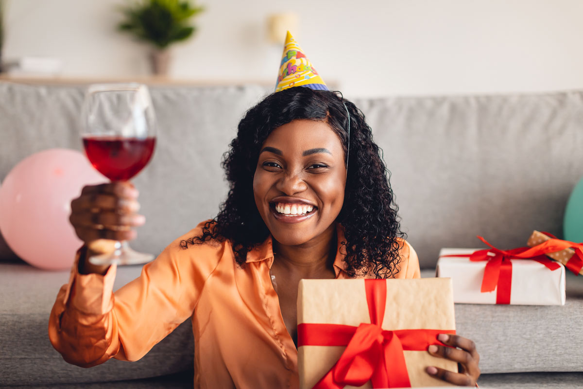 A black 35-year-old woman holding a gold and red present at a birthday party toasts with a glass of red wine.