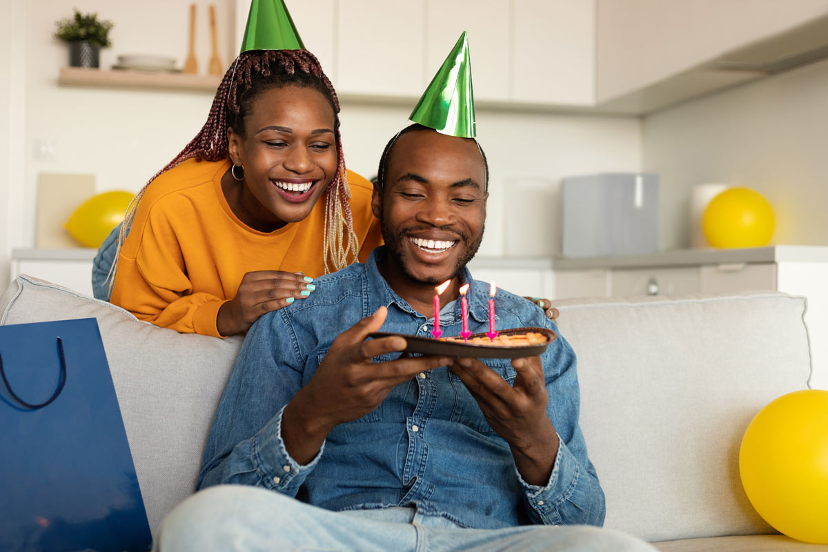 a 30 something black couple smile and laugh while the man sits on a couch surrounded by yellow balloons and holds a pie with 3 candles for his birthday while his partner leans over behind him and grabs his shoulders. 