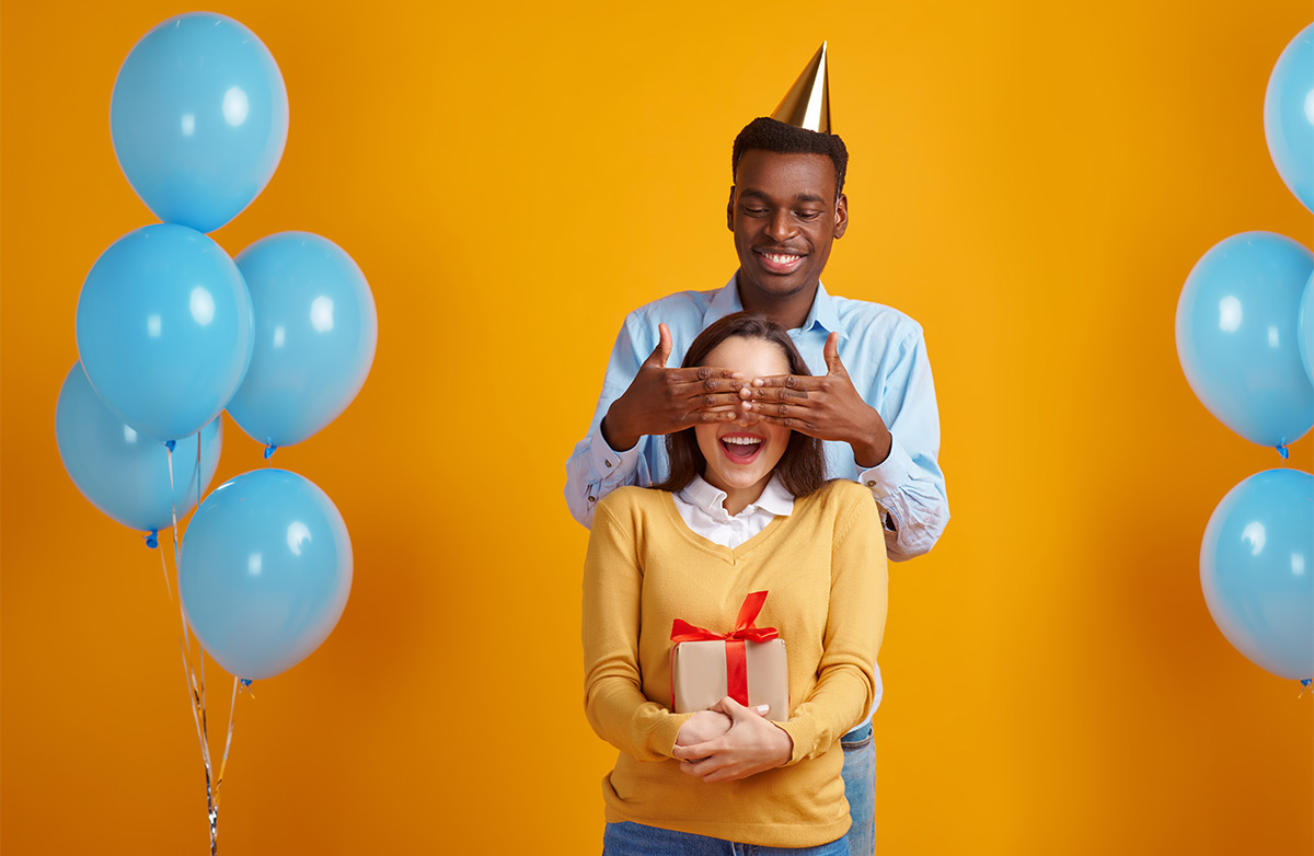 Black man in his 20s-30s covers his white female friend’s eyes for a birthday surprise against a yellow backdrop with blue balloons around them 