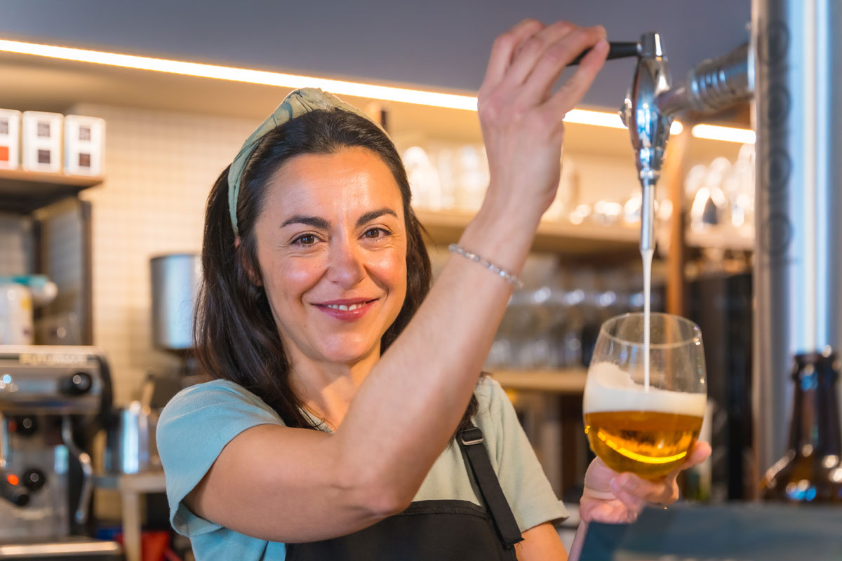 A 35-year-old white woman wearing a green headband smiling at the camera while pouring tap beer. 