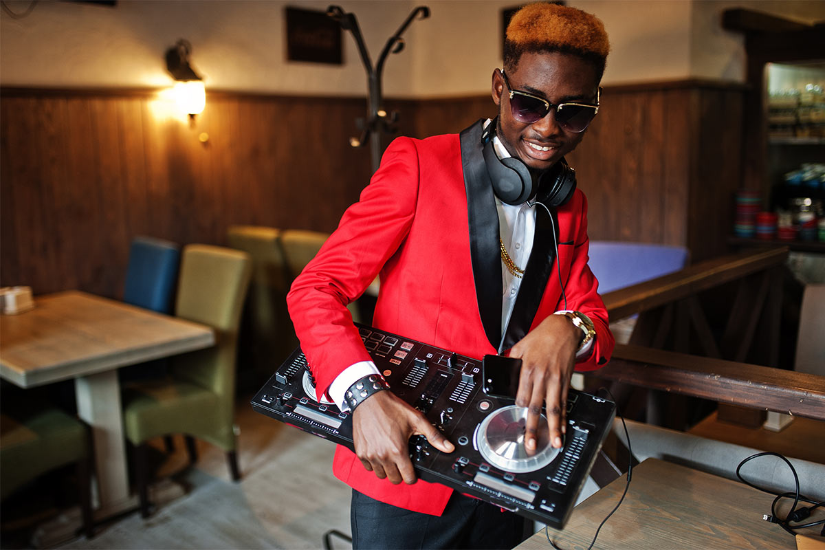 A black teen DJ in a bright red tuxedo and sunglasses, holding a portable turntable and playing music indoors