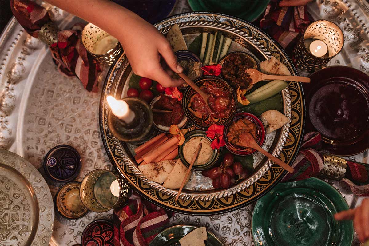 middle eastern woman reaches onto a luxurious platter of middle eastern food that is surrounded by colorful plates, candles, and cloths